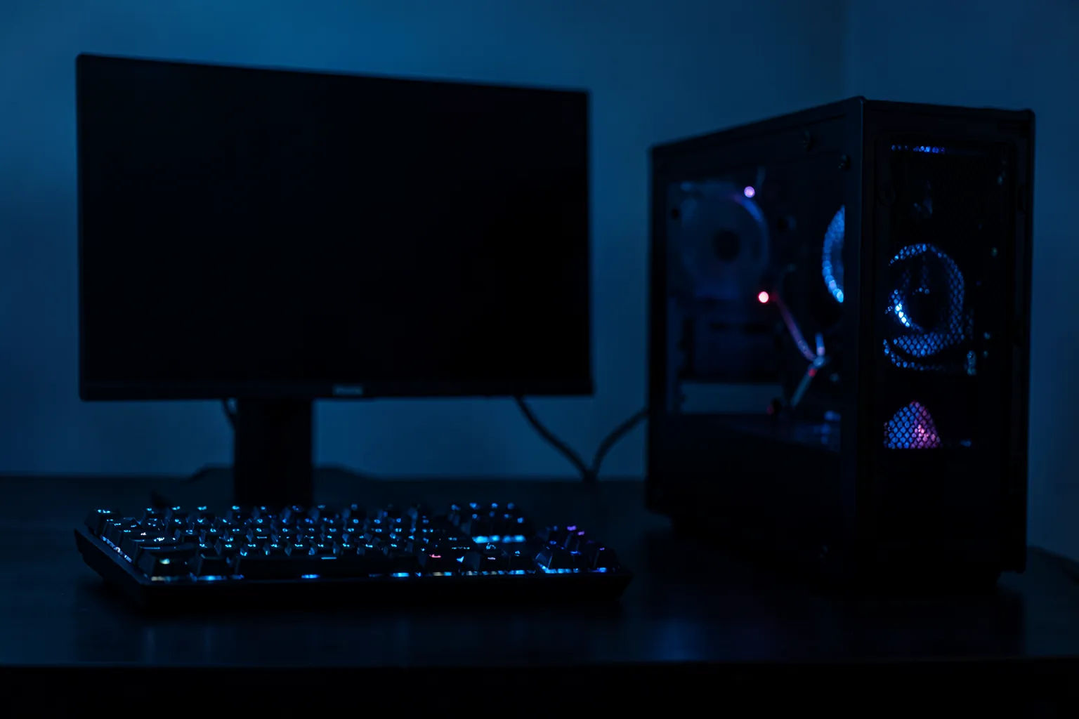 Monitor apagado sobre un escritorio oscuro junto a un teclado mecánico iluminado y una torre de PC con la tapa lateral de cristal