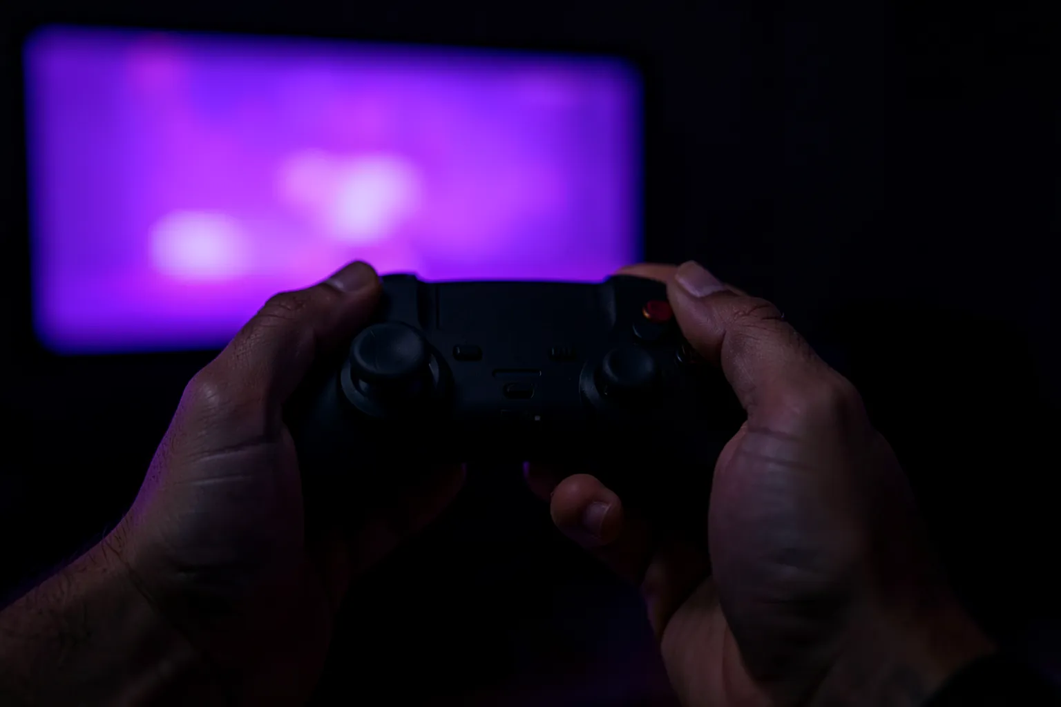Manos sujetando un mando de videojuegos sin marcas frente a un televisor con luz morada, en una habitación oscura