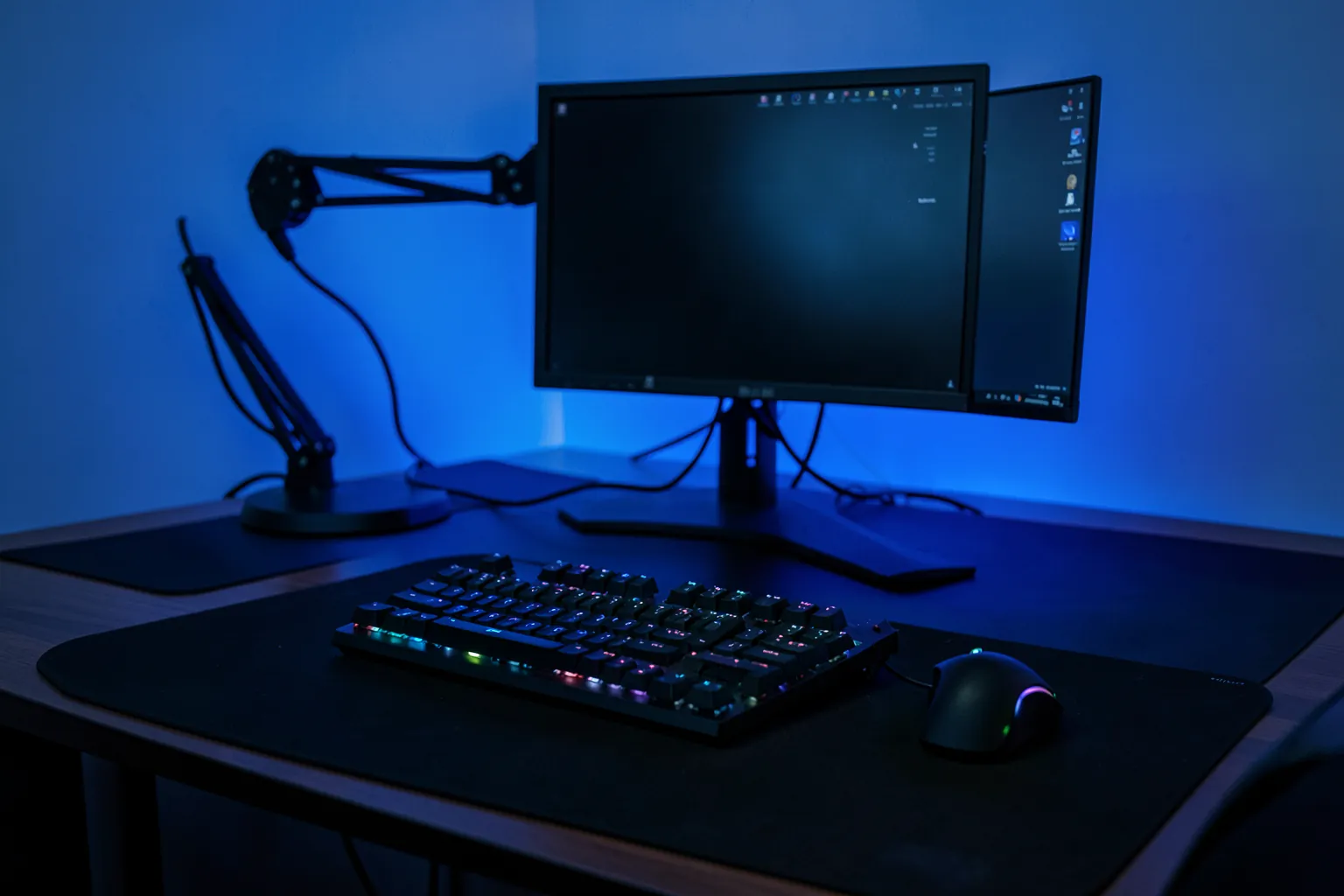 Escritorio gaming ordenado con un monitor en un brazo articulado, un teclado mecánico y un ratón sobre una alfombrilla grande con luz ambiental azul