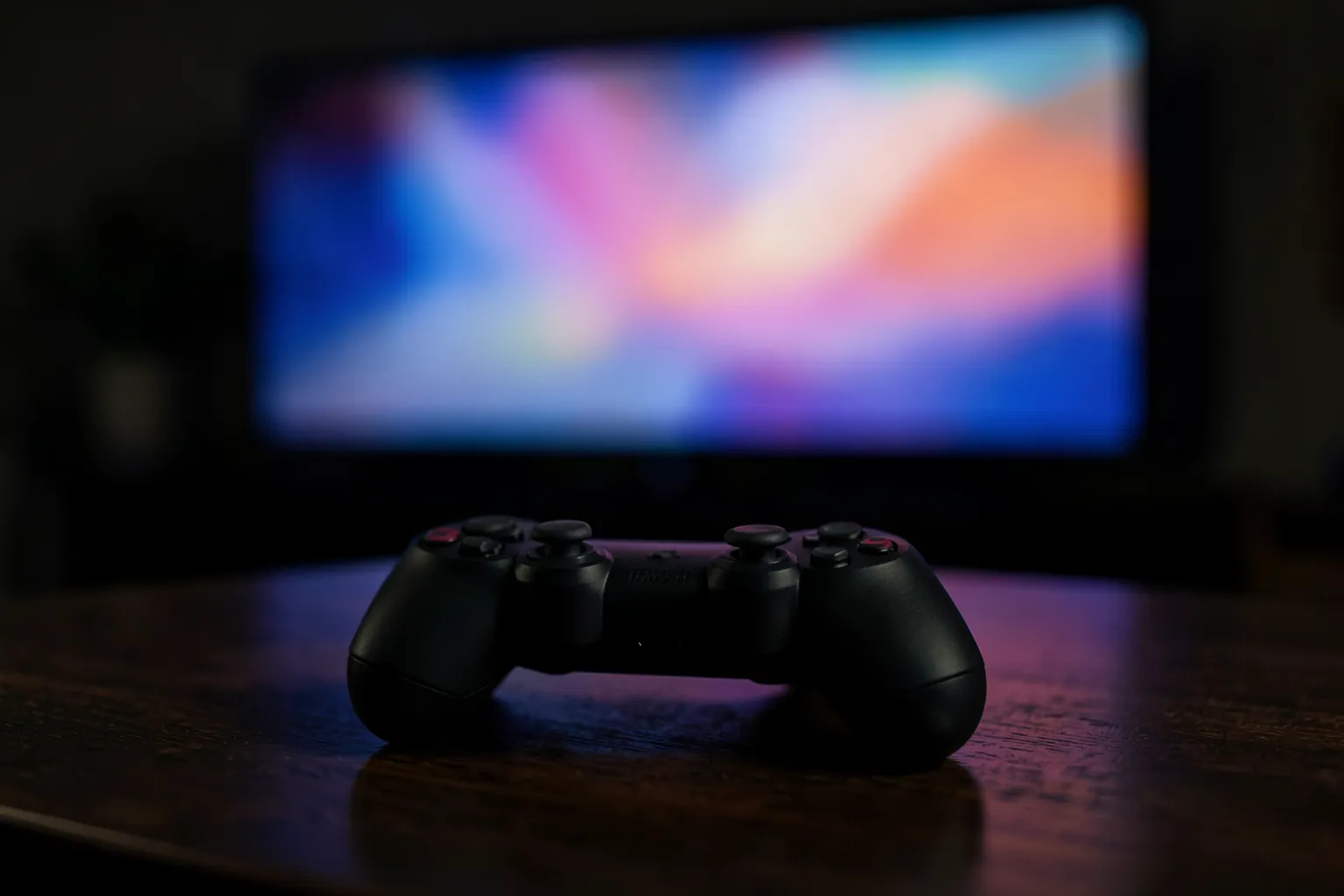 Mando de videojuegos negro sobre una mesa de madera con un televisor desenfocado al fondo que emite colores suaves
