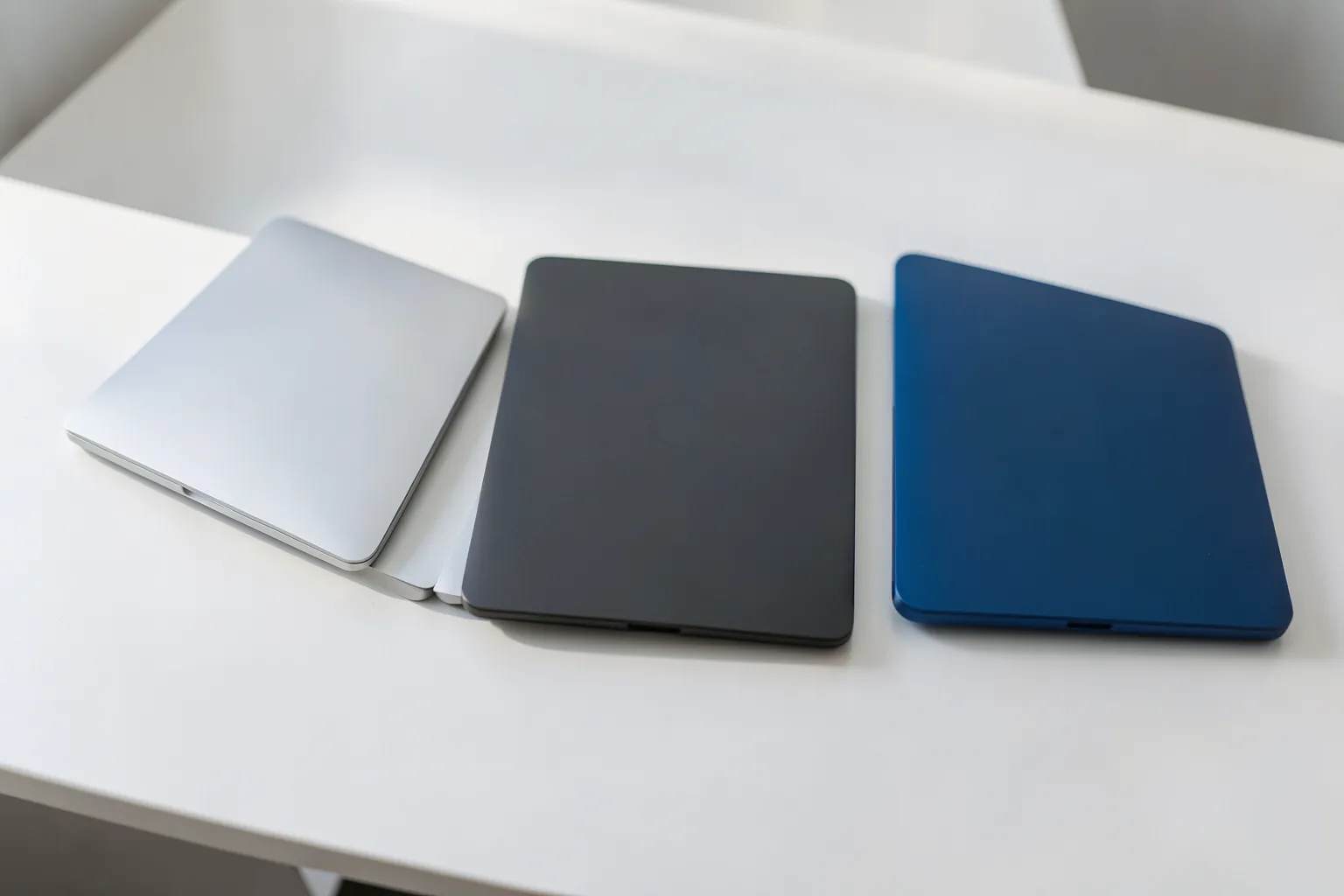 Tres portátiles finos cerrados, plateado, gris oscuro y azul, alineados sobre una mesa blanca con luz natural