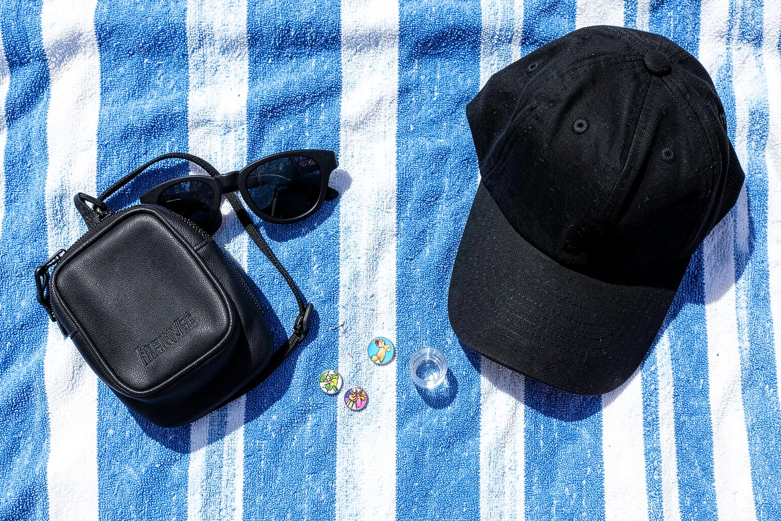 Gafas de sol negras, gorra, riñonera, vaso de chupito y pines de colores ordenados sobre una toalla de playa a rayas