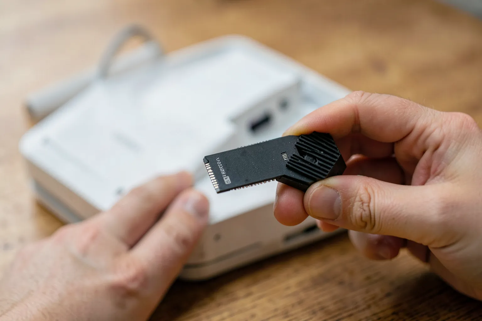Pequeña unidad SSD M.2 con disipador negro en una mano junto a una consola blanca abierta sobre una mesa de madera