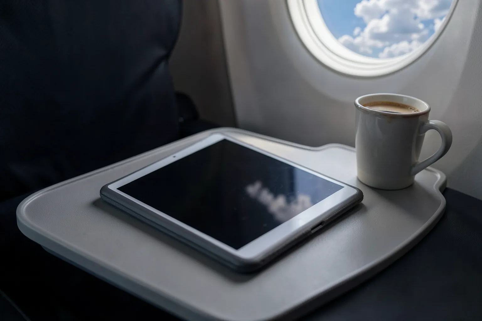 Tableta pequeña con funda gris sobre la bandeja de un asiento de avión junto a una taza de café, con nubes por la ventanilla