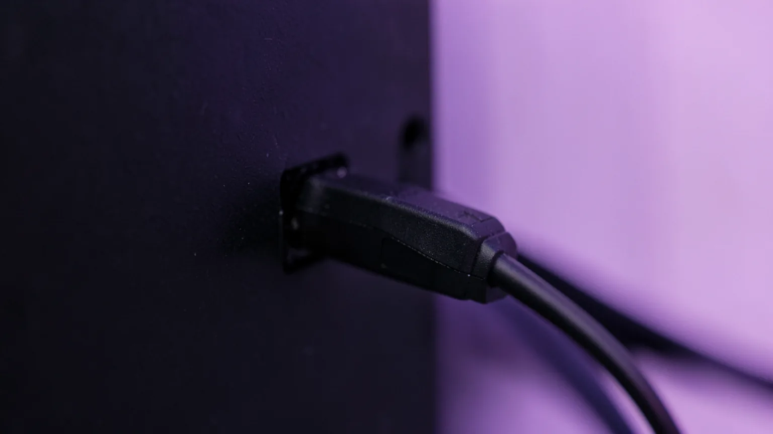 Primer plano de un conector de cable HDMI negro enchufado en el panel trasero de un monitor, con luz violeta de fondo