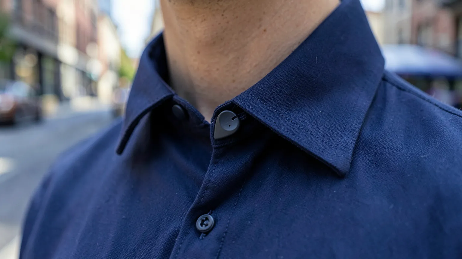 Primer plano del cuello de una camisa azul marino con un diminuto micrófono inalámbrico de solapa enganchado, con una calle desenfocada de fondo