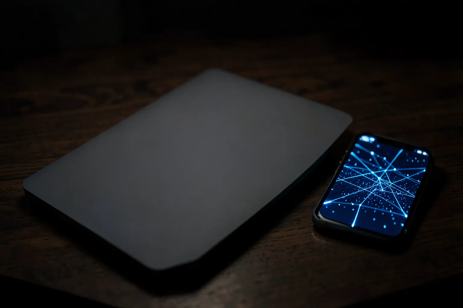 Portátil cerrado sobre una mesa oscura junto a un móvil que muestra una red de líneas luminosas
